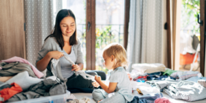 Mom and toddler daughter do chores by folding socks and assorted laundry together on a bed