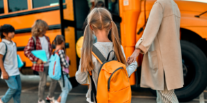 girl with blonde pigtails and a yellow backpack holds her mom's hand as she waits to board the school bus